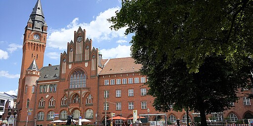  Das Rathaus Köpenick, ein beeindruckendes Gebäude aus rotem Backstein im Stil der norddeutschen Backsteingotik, erhebt sich majestätisch über die Bäume des Luisenhains. Der markante Turm des Rathauses ragt hoch in den Himmel und ist umgeben von üppigem Grün und Wasser, das die Szene friedlich reflektiert. 