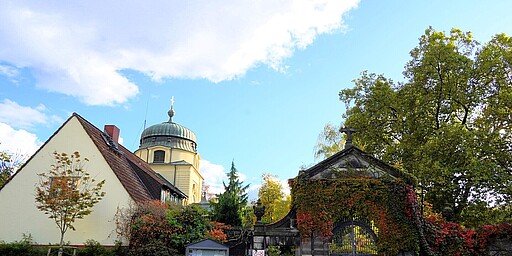 Eingang zum Alten St. Matthäus Kirchhof Der Eingang zum Alten St. Matthäus Kirchhof in Berlin, mit einem Torbogen überwuchert von roten und grünen Blättern. Rechts neben dem Tor ein kleiner Garten und im Hintergrund eine Kuppelkirche mit einem Kreuz an der Spitze.