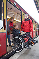 Zwei Hilfskräfte helfen einer Rollstuhlfahrerin beim Aussteigen aus dem Zug