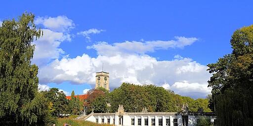 Der historische Pavillon im Rudolph-Wilde-Park  Ein großer Pavillon im Rudolph-Wilde-Park mit einer eleganten weißen Fassade, die sich in einem Teich spiegelt. Im Hintergrund ragt der Turm des Rathaus Schönebergs mit einer Uhr hervor. Der Himmel ist blau, und einige Menschen spazieren durch den Park.