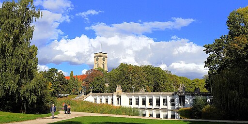  Ein großer Pavillon im Rudolph-Wilde-Park mit einer eleganten weißen Fassade, die sich in einem Teich spiegelt. Im Hintergrund ragt der Turm des Rathaus Schönebergs mit einer Uhr hervor. Der Himmel ist blau, und einige Menschen spazieren durch den Park. 