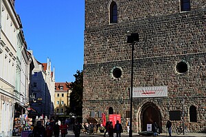 Eine massive Sandsteinmauer der Nikolaikirche im Nikolaiviertel, mit einem großen Eingangstor und runden Fenstern. Passanten schlendern auf der Fußgängerzone entlang, während sich im Hintergrund bunte Gebäude aneinanderreihen. Der Himmel ist klar und blau, und die Szenerie wirkt lebendig.