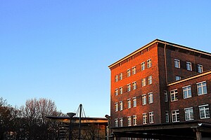 Ein Backsteingebäude, dessen Fassade in einem warmen, rötlichen Licht der untergehenden Sonne glüht. Links im Hintergrund stehen kahle Bäume, daneben ein schlichter moderner Eingang.