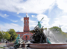 Das Bild zeigt das Rote Rathaus von Berlin mit seinem markanten roten Backsteinbau im Hintergrund. Im Vordergrund sprudelt der Neptunbrunnen mit seinen mythologischen Figuren und Wasserspeiern. Der blaue Himmel ist von weißen Wolken durchzogen.