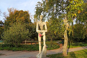 Eine moderne Steinfigur eines nackten Mannes steht auf einem Sockel, umgeben von herbstlichem Grün und Bäumen mit gelben und orangen Blättern. Im Hintergrund ist ein Spazierweg mit einer Person und einem Hund zu sehen. Die Szene wird von warmem Abendlicht beleuchtet.