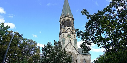Fassade der St. Johannes Basilika Eine imposante Kirche mit einer hohen, spitzen Turmspitze und einer großen Uhr auf der Fassade. Die weiße Fassade ist mit dekorativen gotischen Elementen versehen, und grüne Bäume rahmen das Gebäude ein.