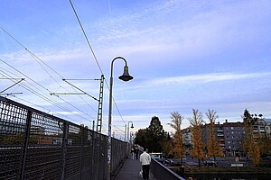 Eine Brücke mit Gehweg, auf der Menschen spazieren. Links das Geländer, rechts herbstlich gefärbte Bäume und ein blauer Himmel.