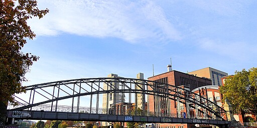 Der Siemenssteg über die Spree mit Blick auf das Heizkraftwerk Charlottenburg  Ein eleganter, gebogener Stahlsteg spannt sich über die Spree. Im Hintergrund ragen die Gebäude und Schornsteine des Heizkraftwerks Charlottenburg empor. Die herbstlich gefärbten Bäume und das klare blaue Himmelsbild schaffen einen Kontrast zur industriellen Kulisse.