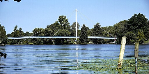  Eine Nahaufnahme des Katzengrabenstegs, die den zentralen Mast und die gespannten Kabel betont. Die Brücke führt über einen breiten Fluss, eingerahmt von grünen Ufern. Im Hintergrund sind vereinzelte Häuser und Bäume zu erkennen. 
