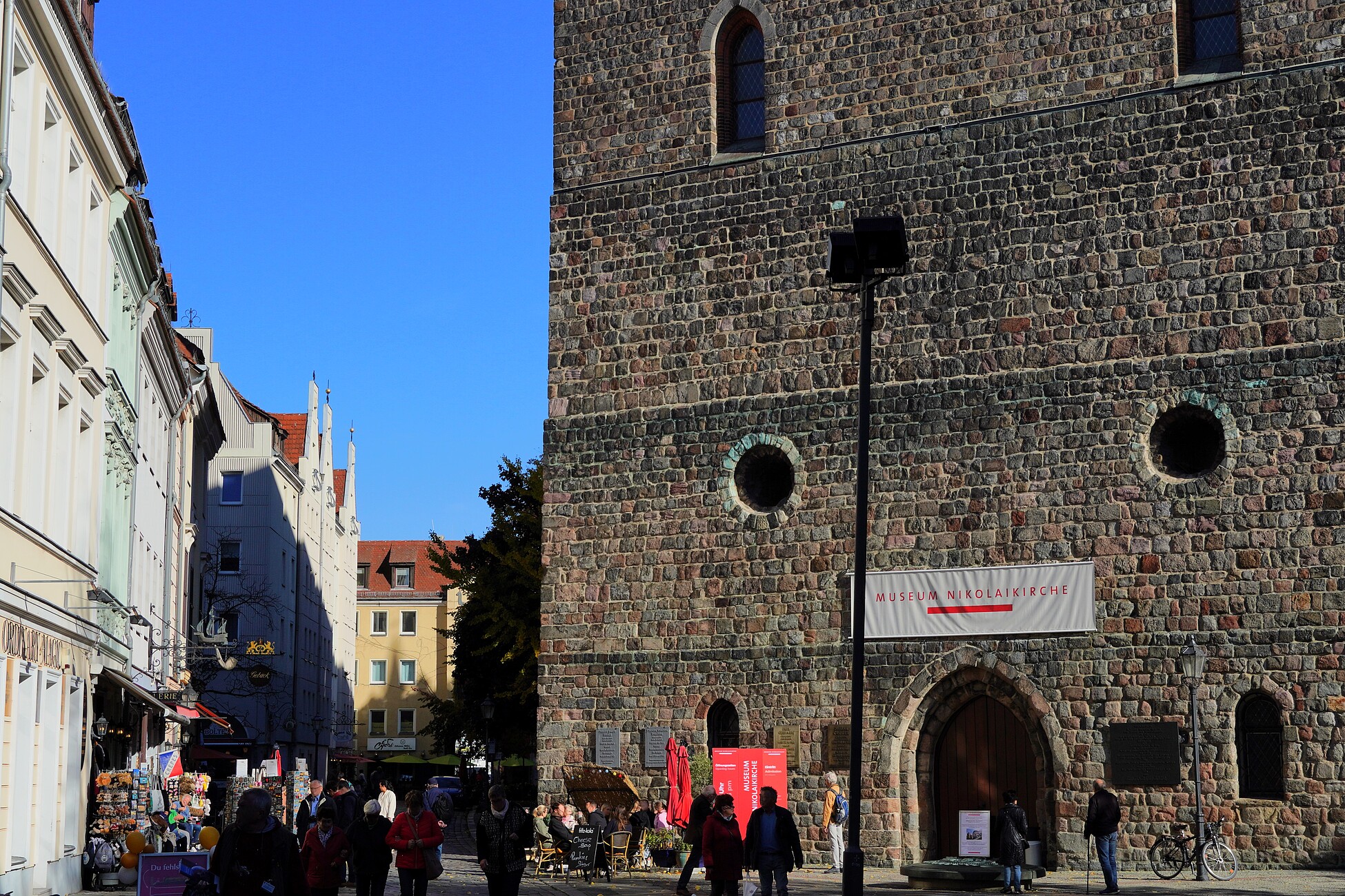 Eine massive Sandsteinmauer der Nikolaikirche im Nikolaiviertel, mit einem großen Eingangstor und runden Fenstern. Passanten schlendern auf der Fußgängerzone entlang, während sich im Hintergrund bunte Gebäude aneinanderreihen. Der Himmel ist klar und blau, und die Szenerie wirkt lebendig.